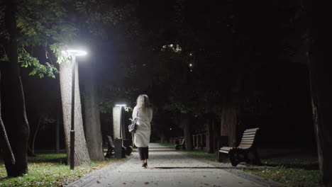 back view of girl with curly hair in white coat carrying black handbag, walking alone down empty park walkway at night, surrounded by benches, trees, fallen leaves and illuminated by streetlamps