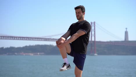 Young-man-in-sportswear-stretching-at-riverside