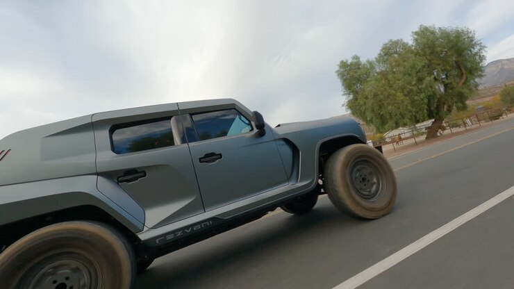 Ripresa FPV con inseguimento di un'auto fuoristrada futuristica che guida su una strada rurale sotto un cielo azzurro