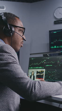 Trader-with-pen-in-hand-works-at-computer-with-displayed-real-time-stocks.-Colleagues-analyze-exchange-market-charts-on-big-screens-at-background.-Office-illuminated-by-blue-light.-Vertical-shot.