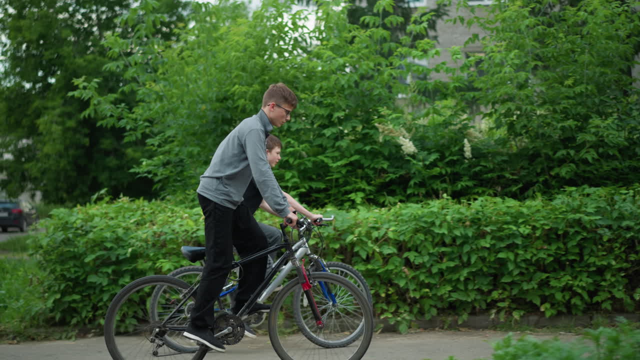 자전거를 타고 있는 두 친구의 측면 전망; 한 명이 회색 을 입고 자전거 타고 서 있고, 다른 한 명이 검은 을 착용하고 자전거에 앉아 있으며, 배경에는 초록색과 흐릿한 건물이 있습니다.