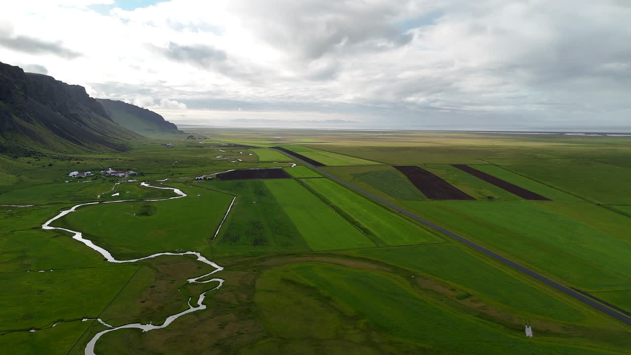 Aerial view of vibrant green fields stretching below steep moss-covered cliffs in Suðurland, Iceland, with winding streams and dramatic shadows shaping the vast volcanic landscape