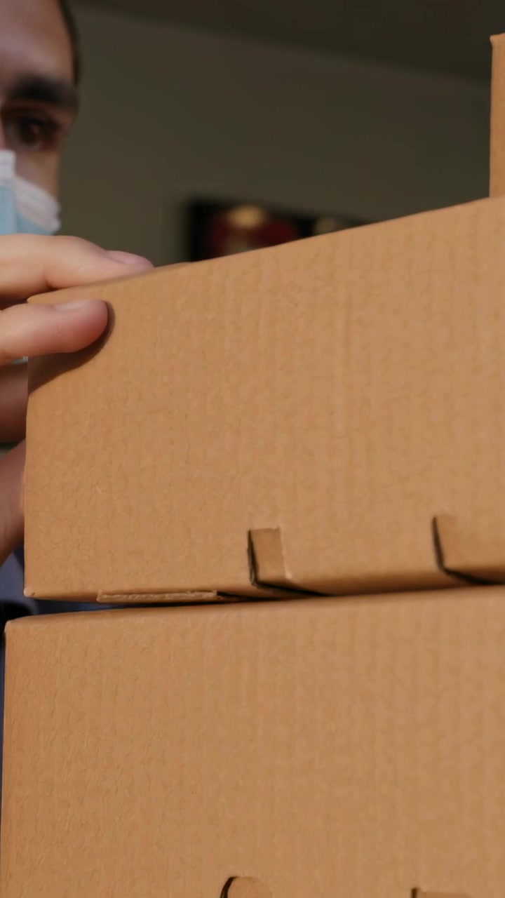 Vertical video: Right-hand reaching worker wearing mask apron steadying 3 boxes in room with hands