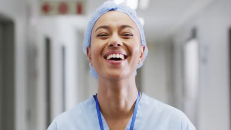 video portrait of happy biracial female medical worker in surgical cap laughing in hospital corridor
