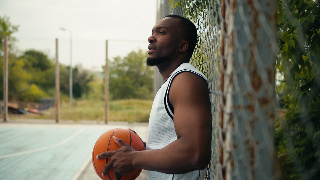 A young black man in a white T-shirt stands near the fence of a basketball court with a basketball in his hands. Active outdoor leisure, sports activities