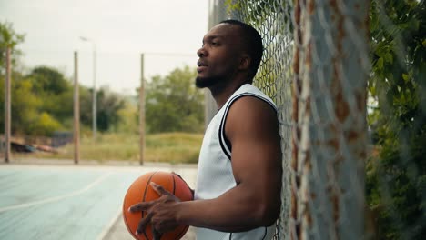 A-young-black-man-in-a-white-T-shirt-stands-near-the-fence-of-a-basketball-court-with-a-basketball-in-his-hands.-Active-outdoor-leisure,-sports-activities