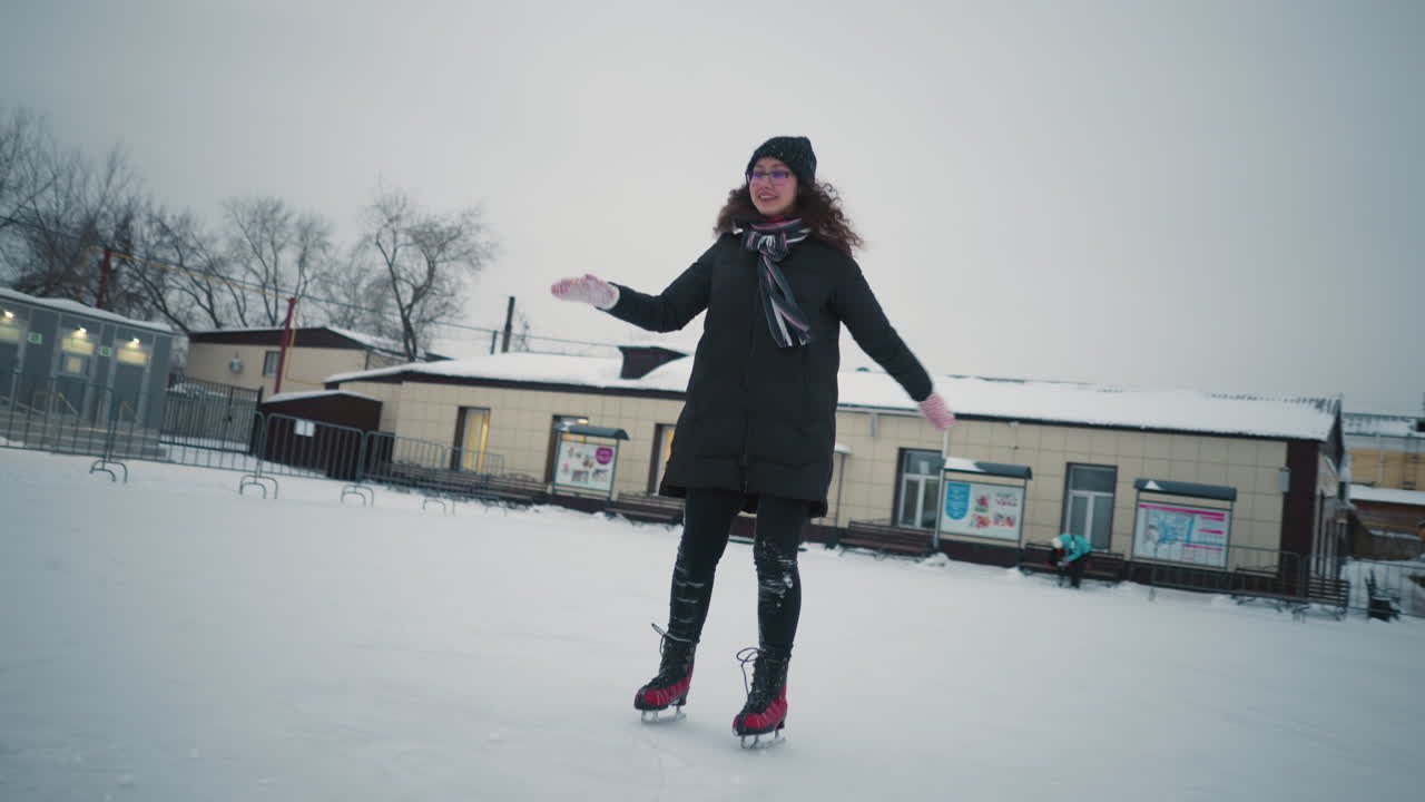 Skater in black winter coat, hat, and red skates gliding across outdoor ice rink with snowy ground, smiling while moving forward, surrounded by benches, small buildings, pine trees, and cold winter sky