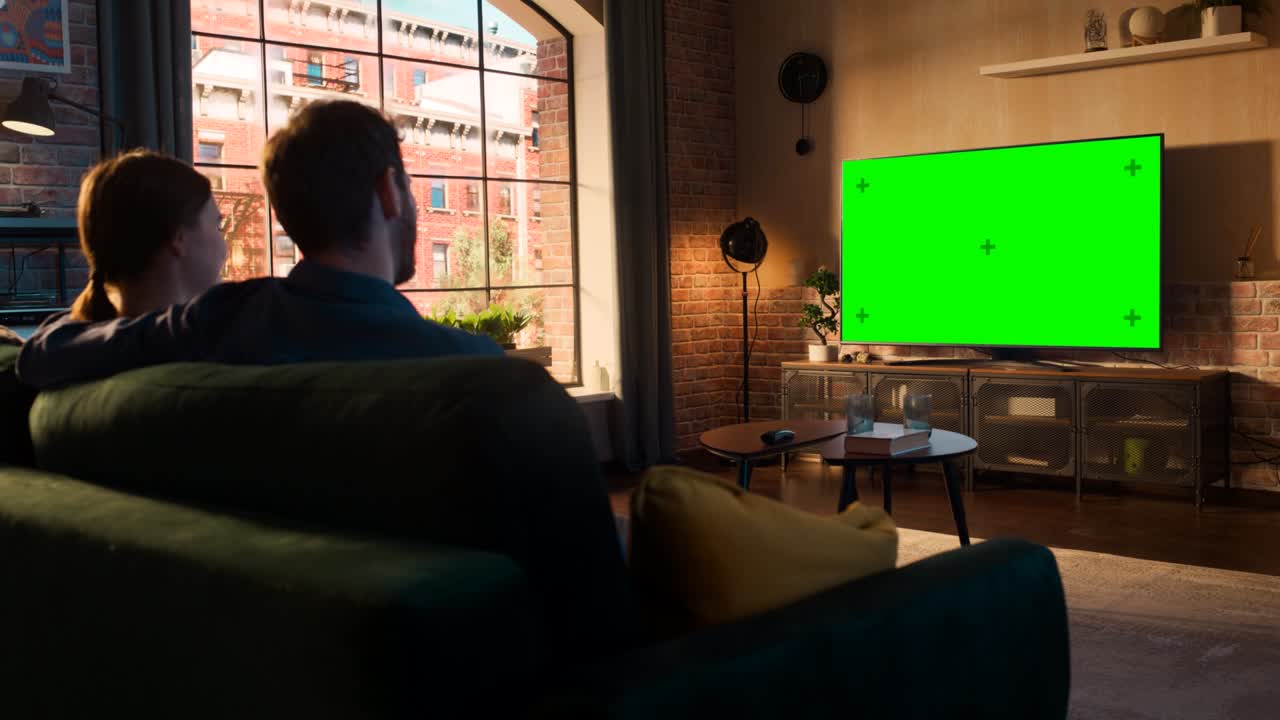 아름다운 부부는 소파에 앉아서 집에서 시간을 보내고, 그들의 세련된 로프트 아파트에서 녹색 화면 모의 디스플레이를 가진 tv를보고 있습니다. 남자와 여자가 영화 또는 쇼를 스트리밍하고 팝콘을 먹습니다.