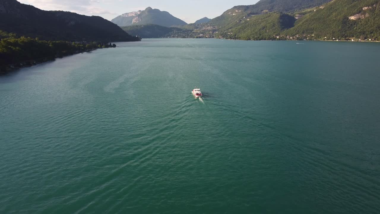 Aerial footage catches up with a ferry travelling along Lake Annecy in France. As the footage catches up with the ferry the drone moves to the right of the ferry as the camera rotates left