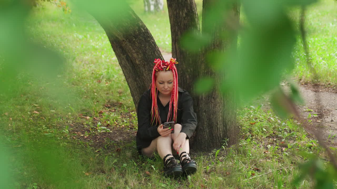 Estudiante caucásico bajo un árbol frondoso enviando mensajes de texto con el teléfono, primer plano bokeh verde, rastas rojas, chaqueta oscura, sentado contra el tronco, pausa reflexiva y tranquila, revisando mensajes, sendero del parque visible, pensativo