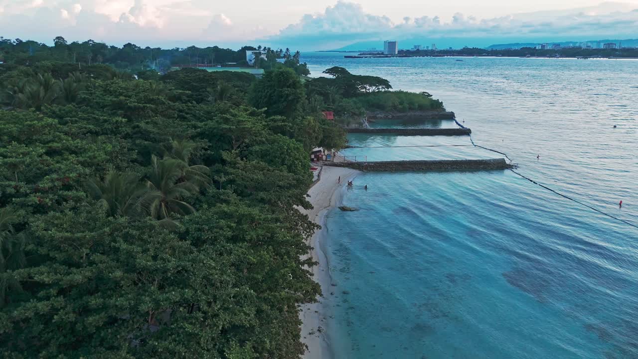 An aerial view of a pristine tropical beach with clear blue water, palm trees, and coastal scenery in the Philippines. The footage captures the serene beauty of nature at sunset or golden hour