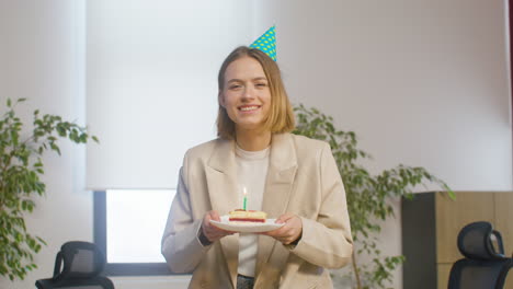 portrait of a happy girl holding a slice of birthday cake on a plate and blowing candle while looking at the camera at the office party