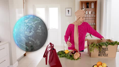 biracial woman in hijab taking vegetables out of box in kitchen over globe