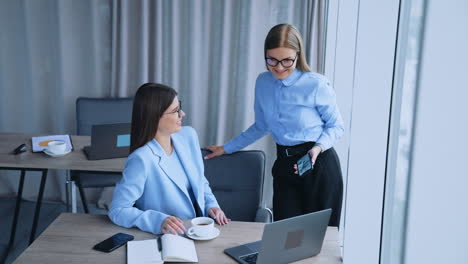female colleagues communication at work. long-haired ladies in glasses talk, laugh and look at laptop. brunette woman having cup of coffee.