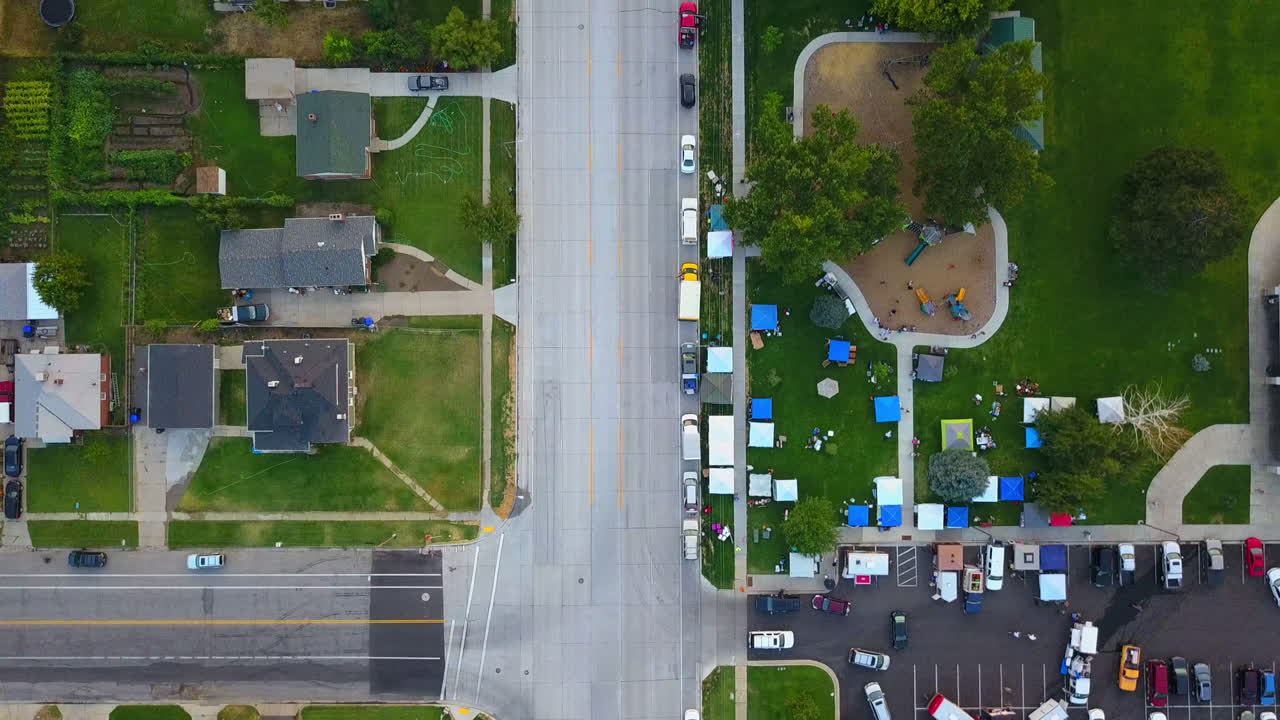A drone shot of a neighborhood street and floating over a park and parking lot next to the park.  The street doesn't have too many cars on it.
