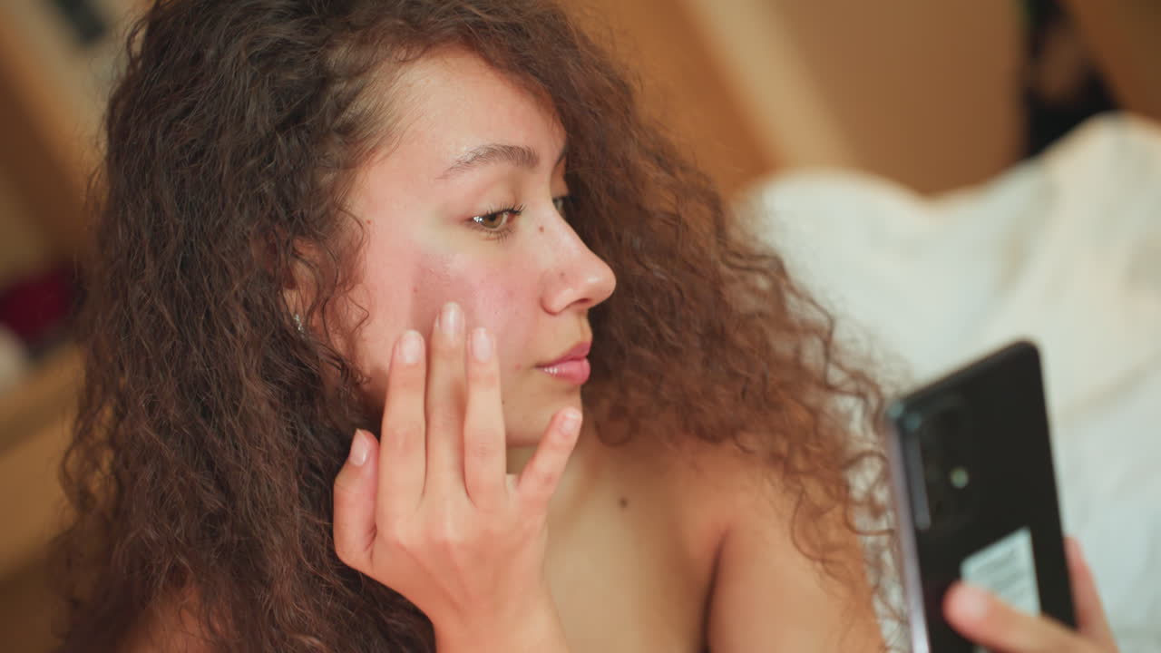 Mulher na cama do hotel pela manhã aplica hidratante usando o espelho do telefone. Iluminação suave e acolhedora, cabelo encaracolado, aplicação de creme com a ponta dos dedos. Um momento de autocuidado focado, de uma hóspede relaxada desfrutando do conforto do hotel.