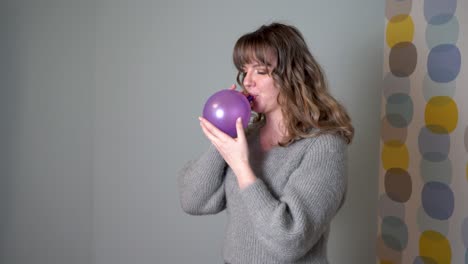 Young-Woman-Blowing-a-Purple-Balloon-In-Front-Of-Grey-Background