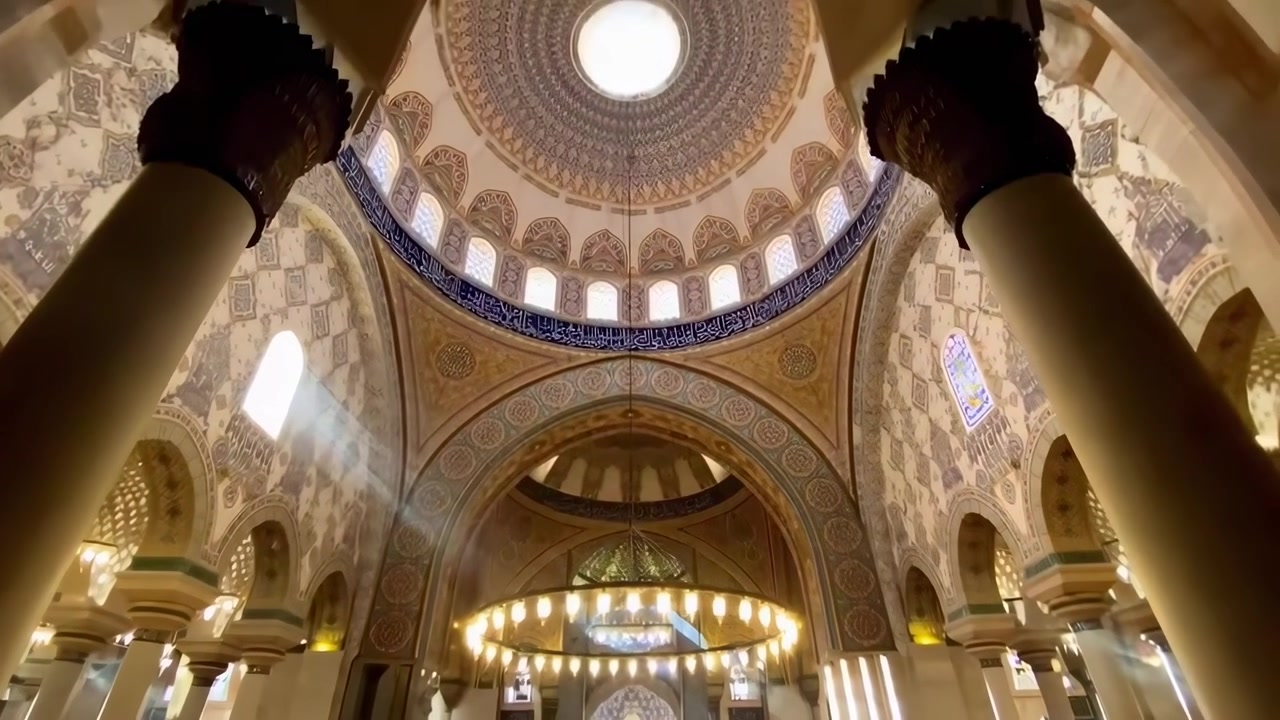Raggi di sole che filtrano nell'interno ornato di una moschea con una grande cupola e un lampadario.