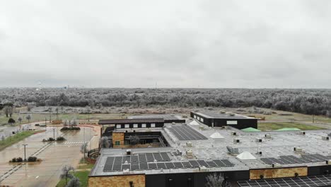 Volando-Hacia-Una-Escuela-Primaria-Que-Tiene-Muchos-Paneles-Solares-En-El-Techo-Y-Un-Enorme-Campo-Y-Una-Sección-De-árboles-Helados-En-La-Distancia.