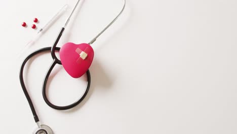 Video-of-red-heart-with-sticking-plasters,-stethoscope,-syringe-and-pills,-on-white-with-copy-space