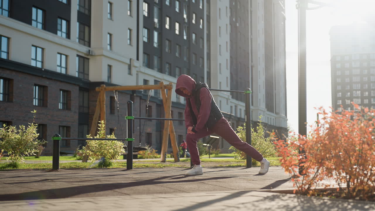 Fitness coach in hoodie and vest at sunset stretches legs forward with hands on bent knee in urban park environment with people walking near playground bars warming up for exercise session