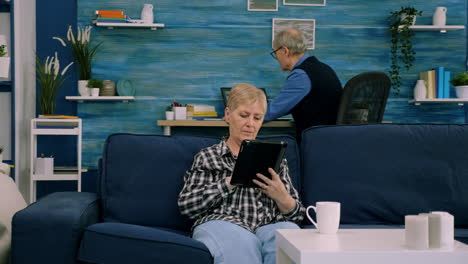 concentrada abuela mayor relajándose sentada en el sofá escribiendo tableta navegando