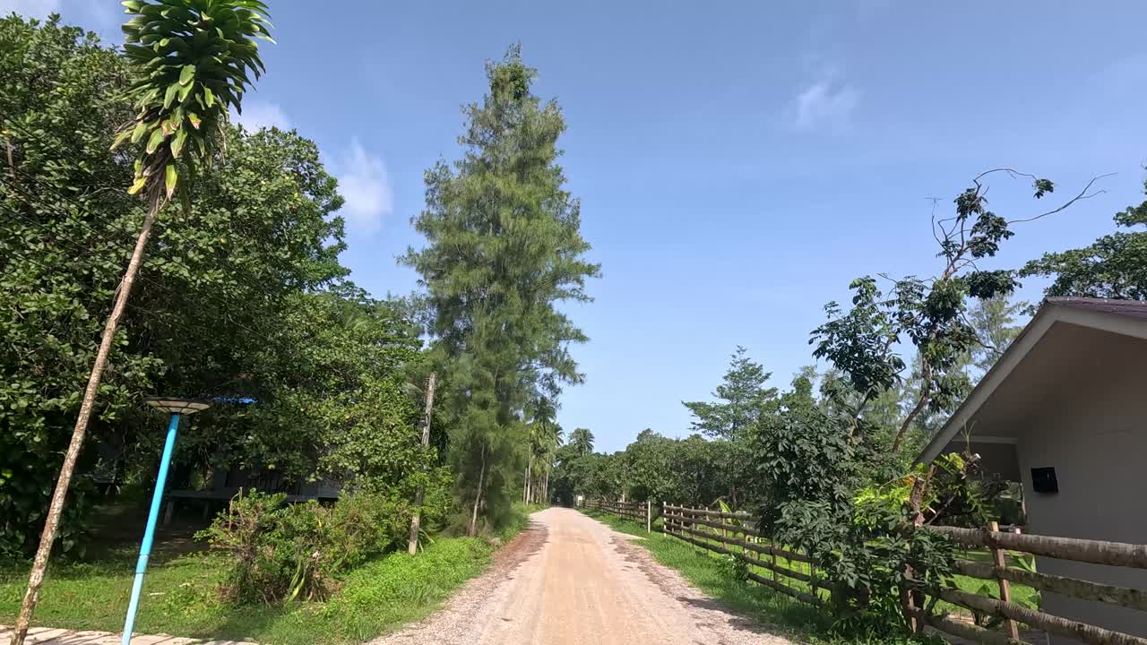 A steady camera moves down a sunlit dirt road lined with houses, lush greenery, and trees on Ko Phayam, Thailand, under clear blue skies