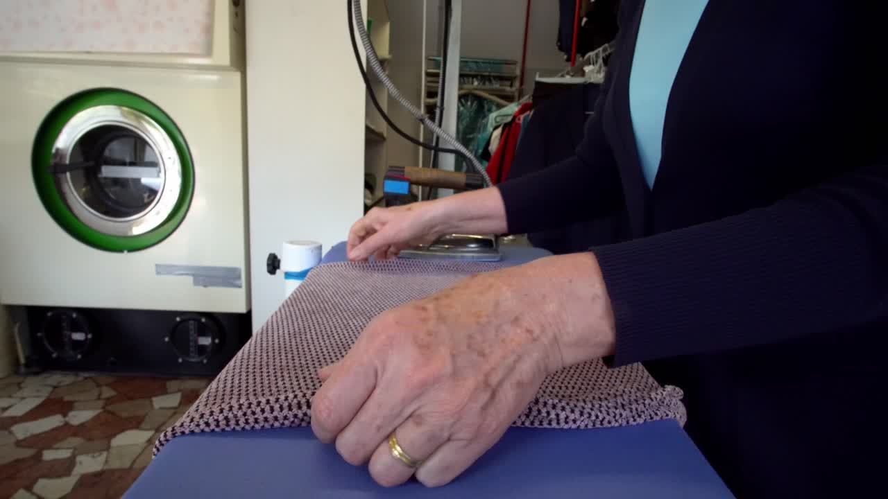 Older woman ironing a folded garment at a laundromat store, locked tripod shot