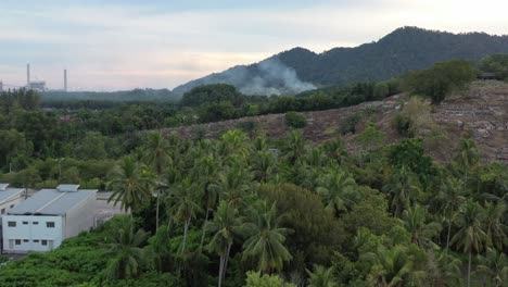 aerial dolly in reveal shot of smoky air pollution produced open air combustion by palm tree plantation industry located next to an active quarry site in district of manjung, perak, southeast asia.