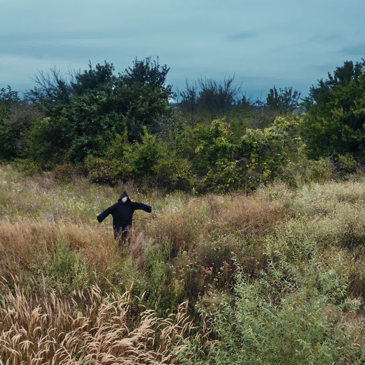 Scary figure of death in the field. Ghost in black cloak standing alone with outstretched arms by sides on nature background.