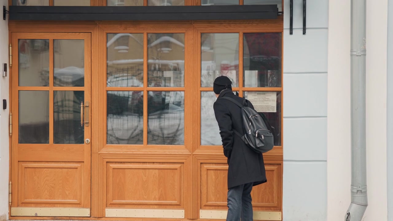Man in black coat and beanie stands outside observing wooden doors with glass panels. Man carries backpack while background shows snow on ground near building exterior. scene is calm