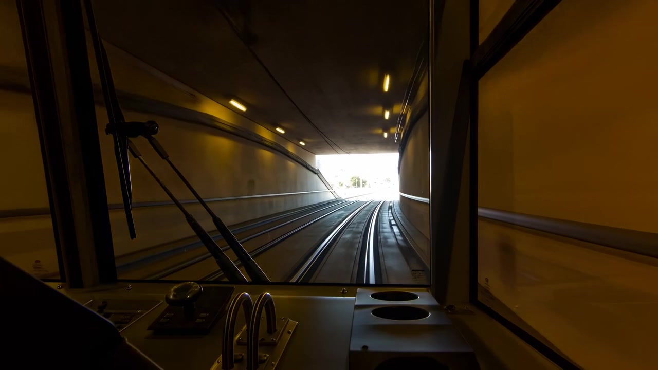 Vista frontale dall'interno di un treno in movimento che entra in una galleria sotterranea fioca, con i binari che curvano in avanti, luci calde che si allontanano in lontananza e una struttura metallica.