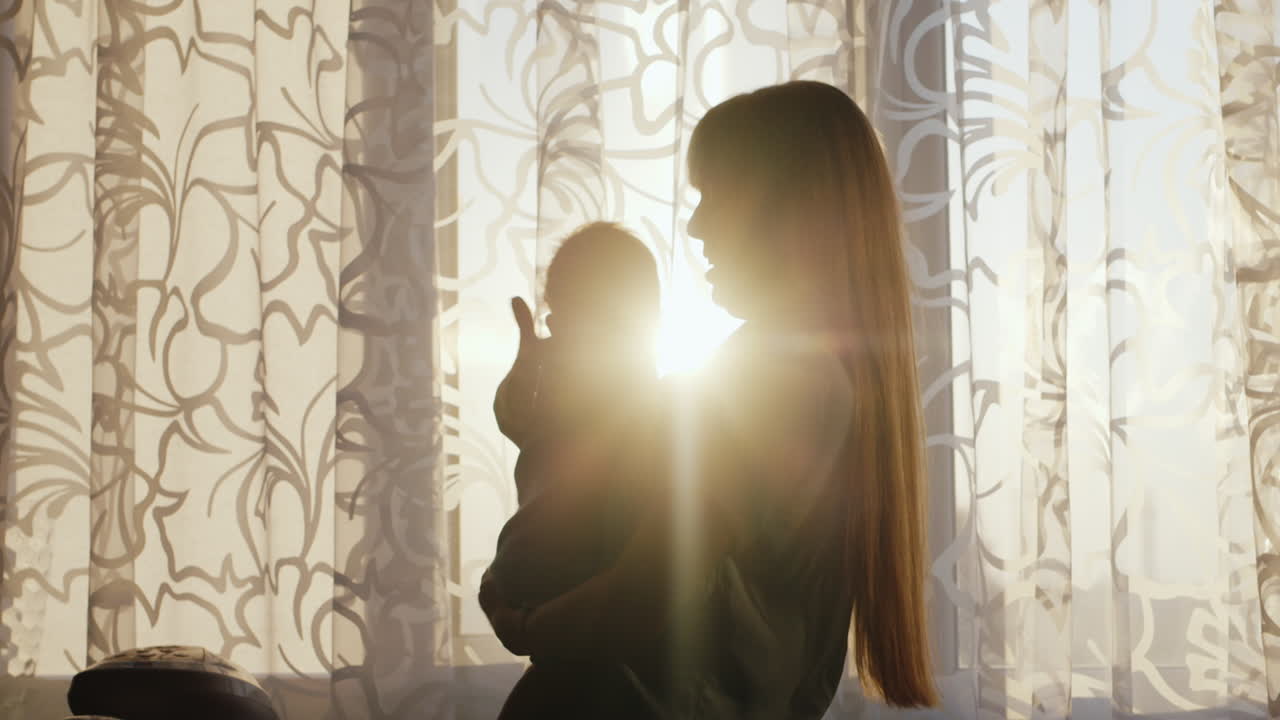 Silhouette Mother With Baby Standing At The Window At Sunset The Sun's Rays Shine Beautifully