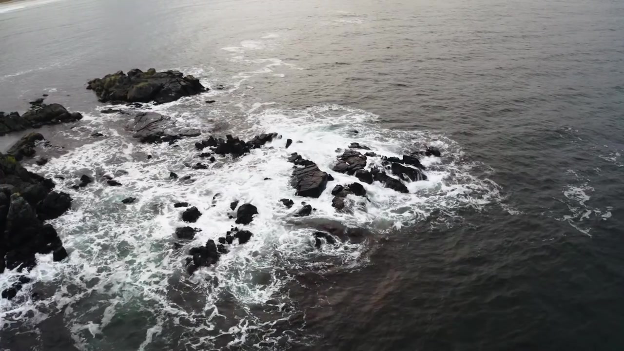 Ampia ripresa aerea in tracking di onde violente che si infrangono su rocce isolate al largo della costa, con una spiaggia e case sul mare sullo sfondo, che mostra la drammatica potenza della costa.