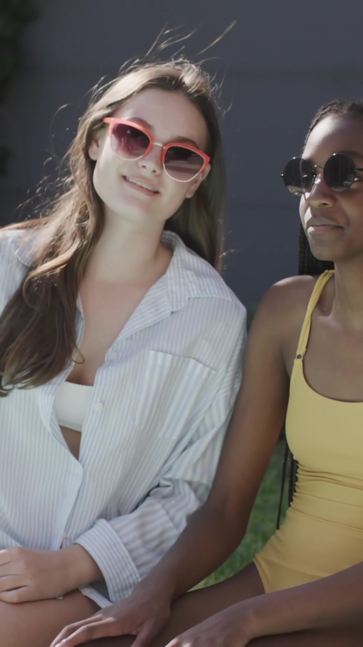 Video verticale: Ragazze adolescenti di diverse etnie in costume da bagno sedute a bordo piscina, che indossano occhiali da sole e avviano una chat al telefono.