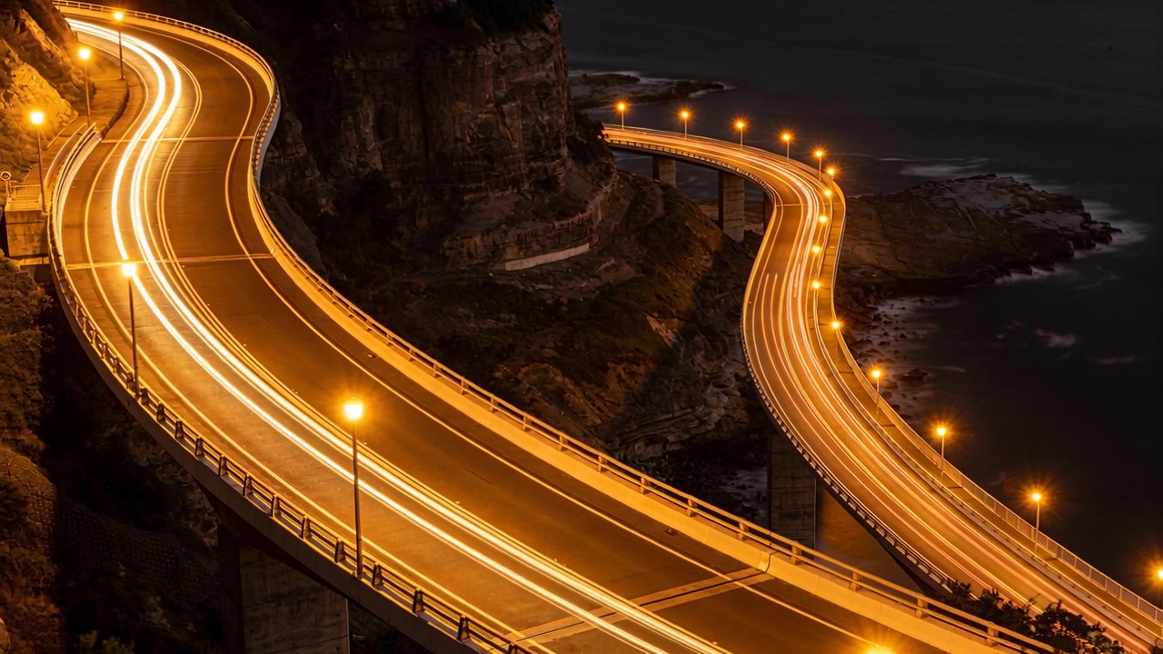 Traffico in movimento lungo due ponti costieri illuminati gemelli che curvano oltre una scogliera di notte, con scie luminose.