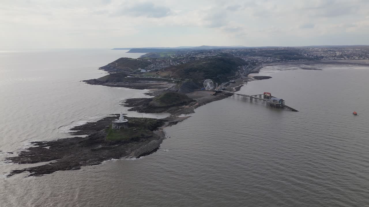 Panoramic aerial orbit with Ferris wheel at Mumbles Pier centered, highlighting the structure against a grey overcast sky near lighthouse