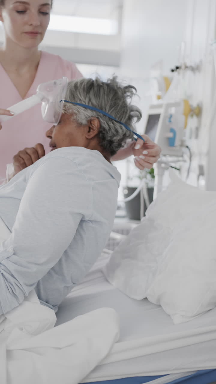 Vertical video of diverse female doctor giving oxygen mask to senior female patient, slow motion