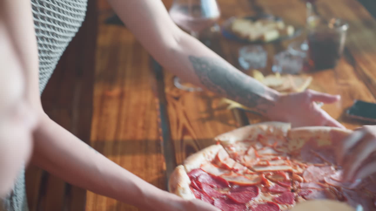 Woman with tattooed hand drops grilled chicken pizza onto table, someone taking a slice to eat, sharing food and enjoying time together in a cozy indoor setting, casual and happy atmosphere