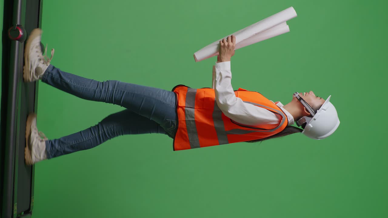 안전 헬을 입은 아시아 여성 엔지니어가 손에 블루프린트를 들고 녹색 스크린 배경 스튜디오에서 는 동안 주위를 둘러보고 있습니다.