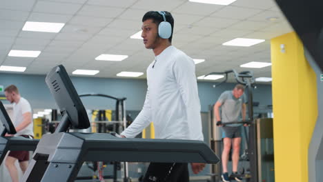 two men walking on treadmills wearing workout gear under bright ceiling lights while third man in background performs dips on parallel bars in spacious gym with modern equipment