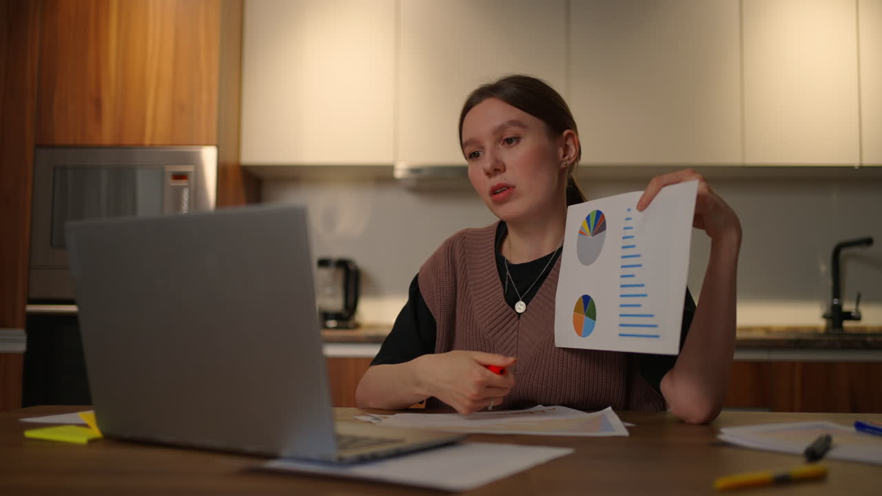 Home office A young woman shows a graph in a laptop camera sitting at home in the kitchen conducting an online report. Video call and conference with demonstration of documents and schedules