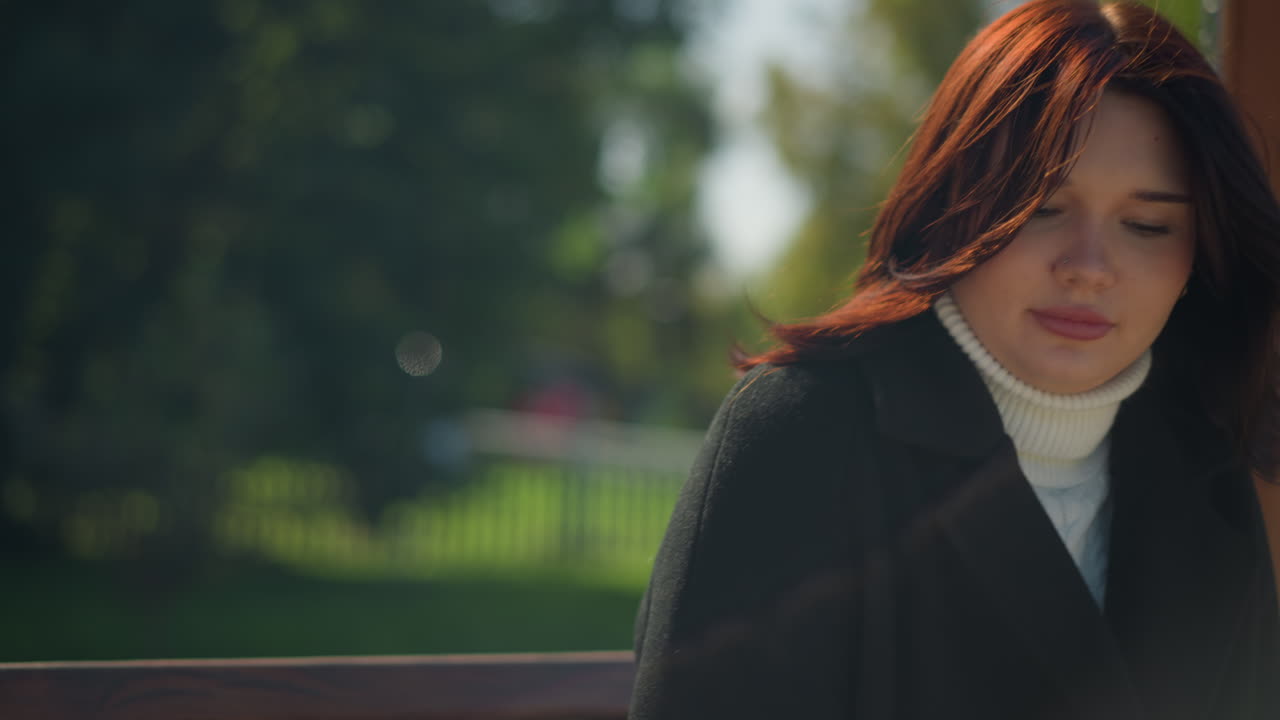 나무로 된 스에 앉아있는 사려 깊은 여자의 클로즈업, 그녀의 머리카락은 부드럽게 바람에 사로잡혔고, 활기찬 초록색 속에서 진정하고 반영적인 분위기를 만니다.