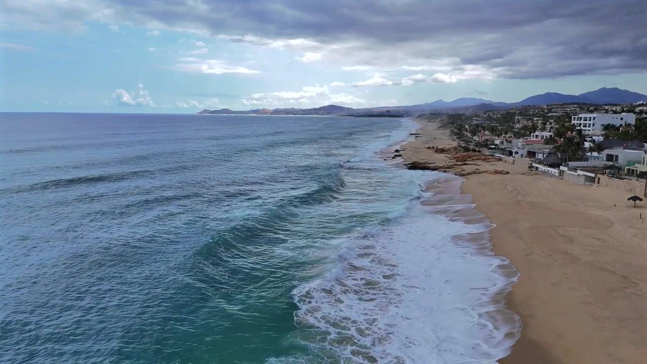 Imagem estática de drone sobre a linha d'água em La Laguna, de frente para Palmilla Point através do oceano, enquanto as ondas quebram na costa, com nuvens de tempestade escuras se formando no interior à direita, ainda fora do enquadramento