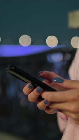 primer plano de una mujer joven con una camiseta rosa sosteniendo un teléfono inteligente, de pie junto a la valla con luces de la ciudad iluminadas y un puente borroso en el fondo por la noche