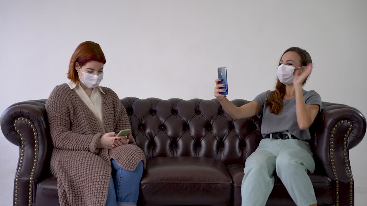 концепция социального дистанцирования. две женщины в медицинских масках, разговаривают на смартфоне, делают селфи, сидят на диване.
