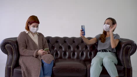 사회적 거리두기 개념. 의료용 얼굴 마스크를 착용한 두 명의 여성이 스마트폰으로 채팅하고, 셀카를 찍고, 소파에 앉아 있습니다.