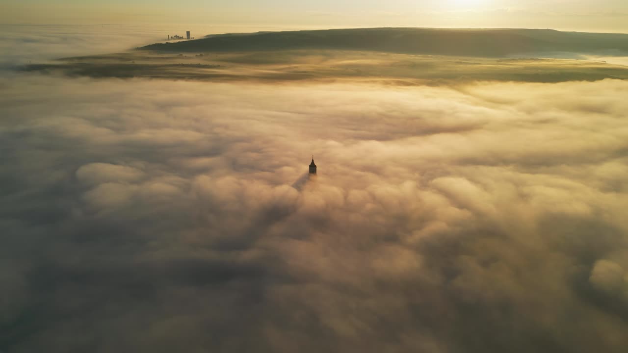 Italy Matera, drone video orbiting around partially visible tower that protrudes through low hanging clouds during sunrise, golden colours incredible view