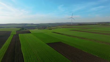 Wind-turbine-stands-tall-amidst-vast-green-fields-under-a-clear-blue-sky,-aerial-view