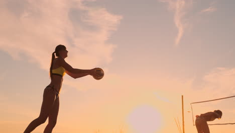 Eine-Schöne-Frau-Im-Bikini-Mit-Einem-Ball-Macht-Sich-Bei-Sonnenuntergang-Bereit,-Bei-Einem-Volleyballspiel-Im-Sand-Am-Strand-Einen-Aufschlagsprung-Zu-Machen.-Der-Entscheidende-Moment,-Der-Spannende-Moment-Des-Spiels-In-Zeitlupe.